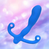 The Aneros Helix Syn V Special Edition Blue, a curved vibrating prostate massager with two looped handles, is displayed against a soft pastel pink and purple bokeh background.